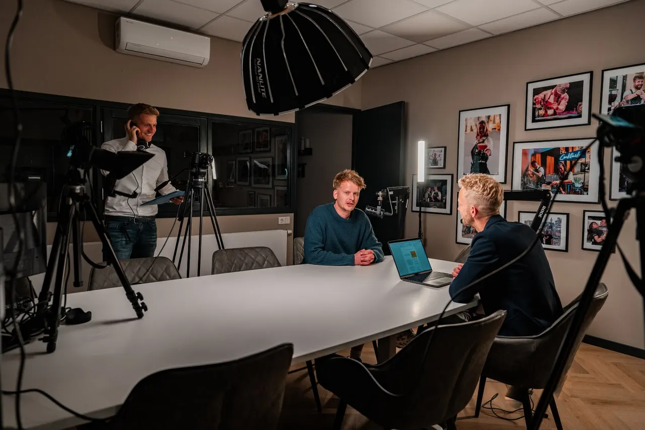Vergaderruimte omgebouwd tot studio: camera's op statief, een softbox boven de tafel en een crewlid met koptelefoon tijdens de opname.