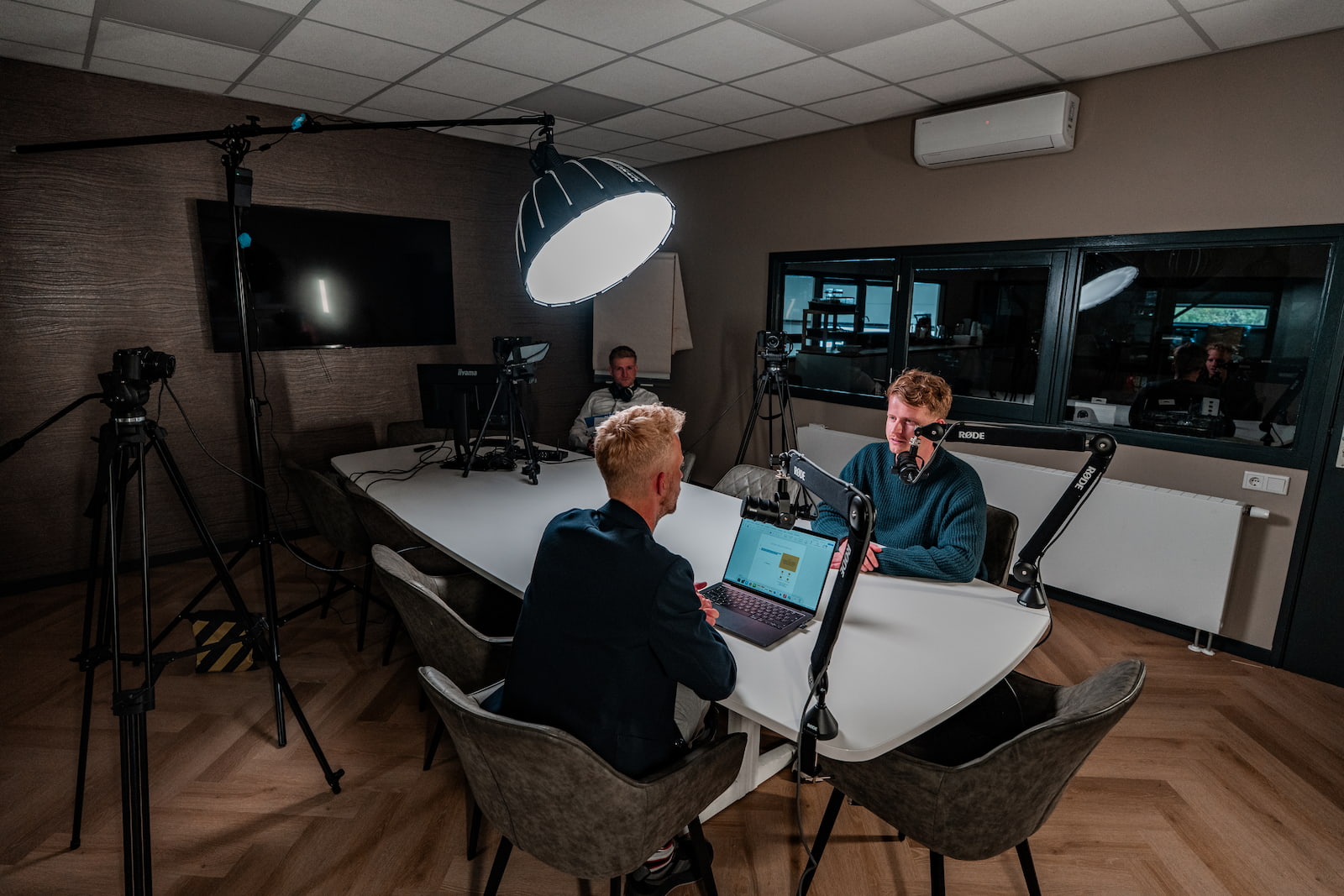 Opnamesessie met twee hosts en een gast aan de studiotafel, met boom-microfoons en camera.